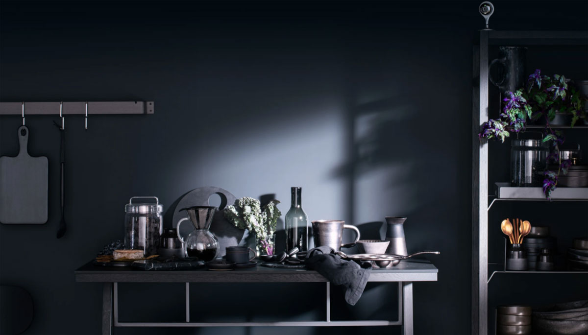   In the center of picture are some glass wares on a desk, on the right part of the picture is a shelf with some kitchen ware and potted place 